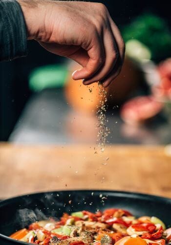 Hand of person sprinkling dried oregano in a frying pan with veggies